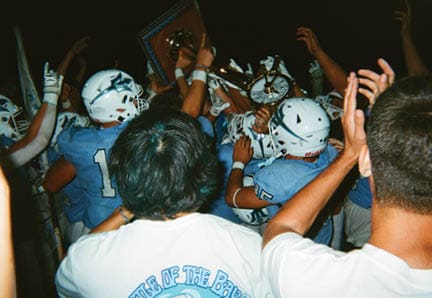 A group of people wearing black football helmets and jerseys are gathered closely together on a field. Description generated by AI
