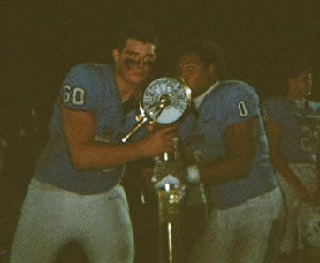 A group of people wearing black football uniforms with “VISTA" on the front are celebrating energetically on a field during daylight. Description generated by AI