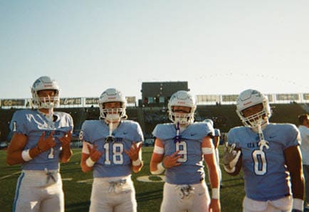 Four people in football uniforms are positioned on a field, appearing ready to start a play during a game. Description generated by AI