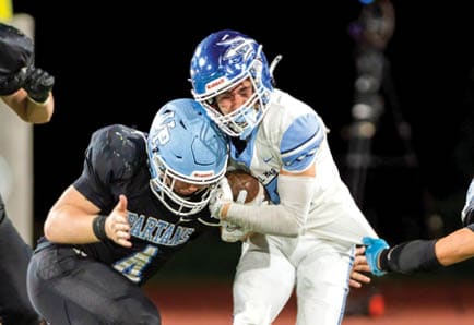 Two adults in football uniforms are actively engaged in a play on a field, with one holding the ball and the other preparing to tackle. Description generated by AI