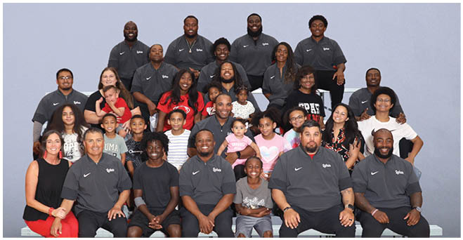 A large group of people and children are posed together against a plain background, with many wearing matching dark gray polo shirts and smiling. Description generated by AI
