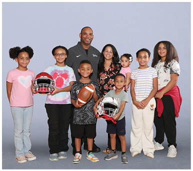 Two children smiling with one holding a football and the other holding a football helmet, standing next to an adult wearing a floral patterned shirt. Description generated by AI