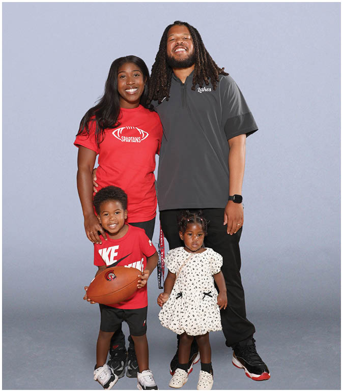 A group of four people, including two adults and two children, stand together smiling with one child holding a football. Description generated by AI