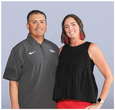 Two adults stand side by side against a plain background, one wearing a dark gray Nike pullover and the other wearing a sleeveless black top with red pants and red earrings. Description generated by AI