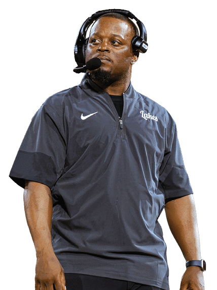A person wearing a dark Nike shirt and pants with a headset and microphone stands on a sports field. Description generated by AI