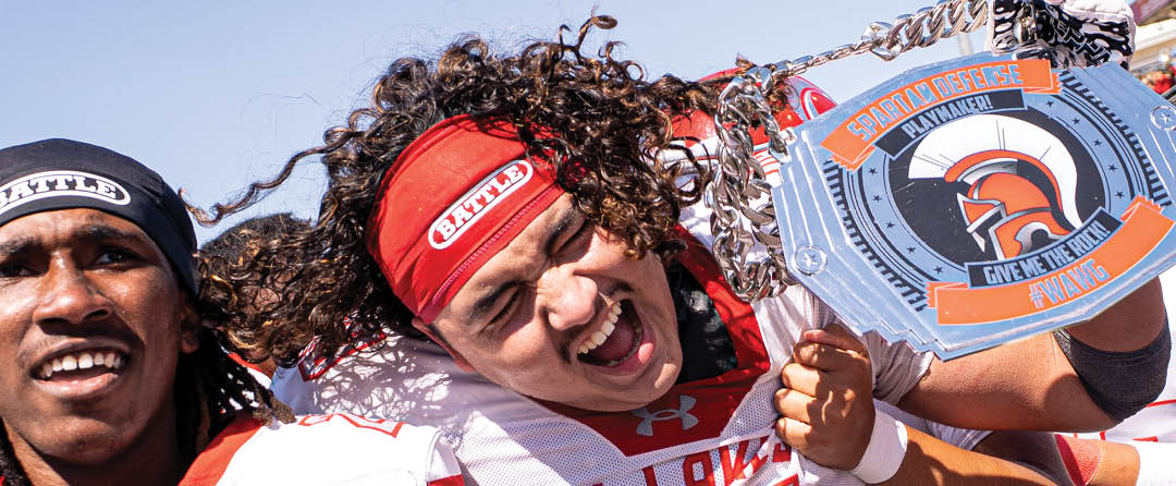 A person with curly hair and a red headband is holding a large circular sign with the words “SPARTAN OBSTACLE PLAYMAKER" outdoors under a clear sky. Description generated by AI