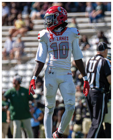 A person wearing a red football helmet, white jersey with red lettering and the number 10, white pants, and red gloves stands on a football field with blurred spectators in the background. Description generated by AI