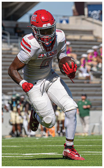A person wearing a red helmet and white football uniform with red accents is running while holding a football on a field. Description generated by AI