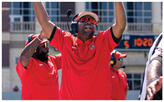 A person wearing a red sports shirt, headset, and sunglasses is raising their arms enthusiastically while another person in similar attire looks on in the background. Description generated by AI