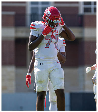 A person wearing a red helmet and white football uniform with the number 71 is adjusting their helmet on a sports field. Description generated by AI