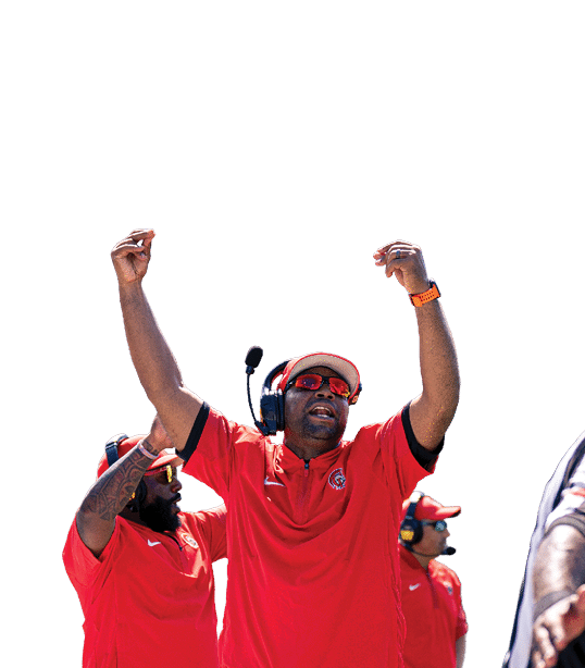 A person wearing a red shirt, headset, and sunglasses is raising both arms in celebration on a sports field with other people in red shirts and a scoreboard in the background. Description generated by AI