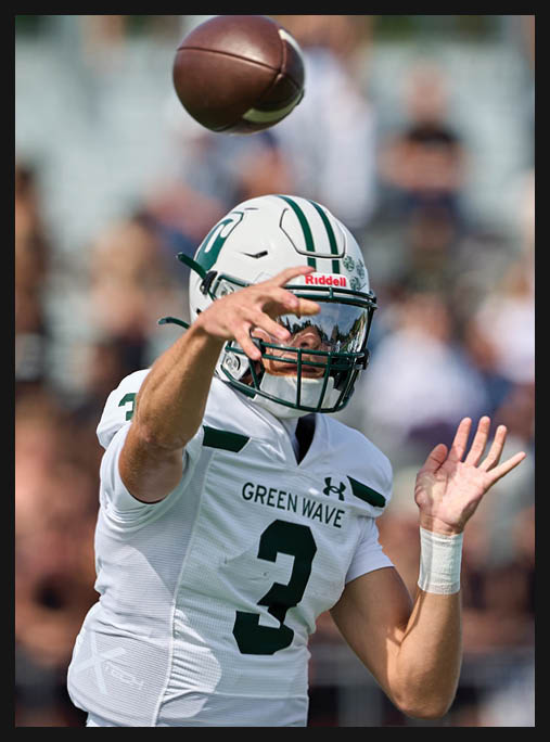 Two people in green football uniforms kneel on a field holding their helmets with heads bowed while cheerleaders stand blurred in the background. Description generated by AI