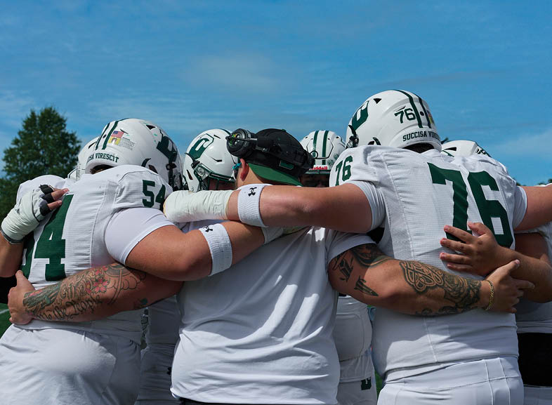 Four people wearing black football uniforms with “VISTA" on the front are standing together in a stadium with a crowd in the background, two of them holding helmets raised above their heads. Description generated by AI