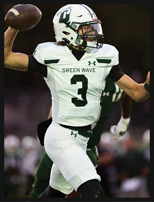 Two people in green football uniforms kneel on a field holding their helmets with heads bowed while cheerleaders stand blurred in the background. Description generated by AI
