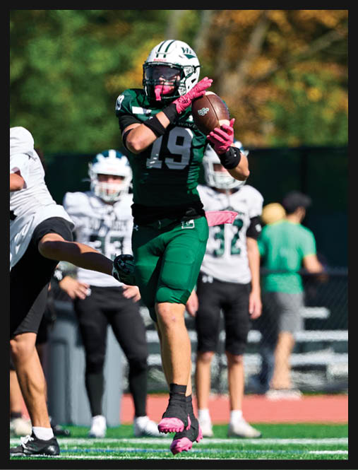 Two people in green football uniforms kneel on a field holding their helmets with heads bowed while cheerleaders stand blurred in the background. Description generated by AI