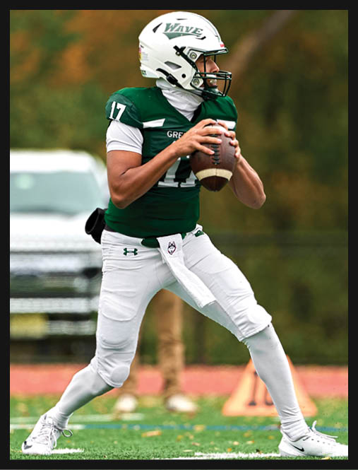 Two people in green football uniforms kneel on a field holding their helmets with heads bowed while cheerleaders stand blurred in the background. Description generated by AI