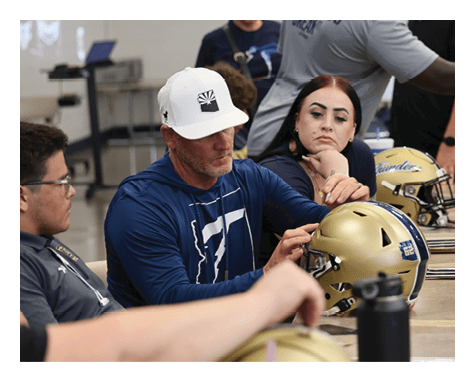 A group of people are gathered around a table with football helmets on it. A man is holding a football helmet while a woman looks on. There are several other people in the room, some of whom are wearing backpacks. A bottle is also visible on the table. AI generated content
