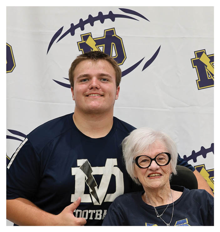 A man and woman pose together for a photo. The woman is wearing a blue shirt and glasses, while the man is wearing a blue shirt and a football helmet. AI generated content