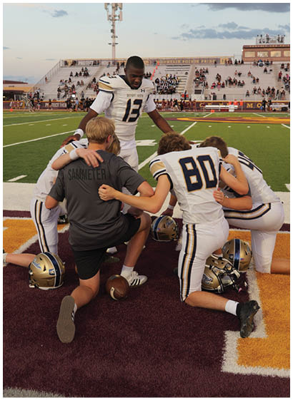 A group of football players and a coach kneel together on the field in a huddle with helmets on the ground nearby. Description generated by AI