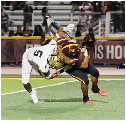 Two people in football uniforms are engaged in a tackle on a field with one person lifting the other off the ground. Description generated by AI