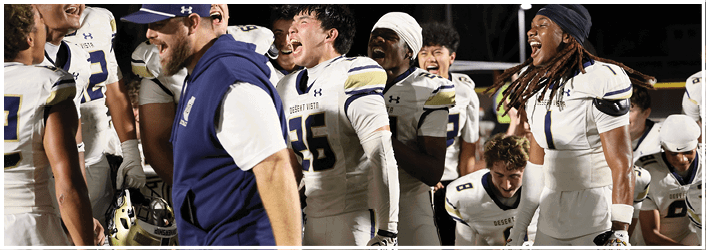 People in football uniforms are gathered closely on the field, some holding helmets and one person wearing a blue vest standing among them. Description generated by AI