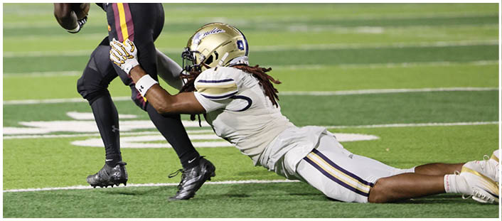 A person in a white football uniform is diving and grabbing the leg of another person in a black football uniform who is running on a field. Description generated by AI