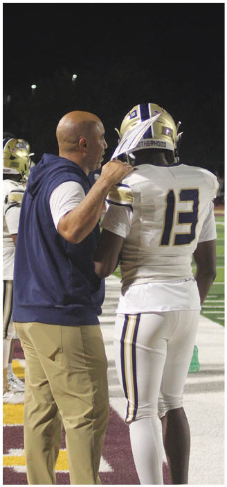 An adult wearing a sleeveless hoodie is talking to a person in a football uniform with the number 13 on the field during a nighttime game. Description generated by AI