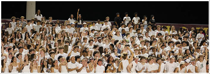 A large group of people wearing white shirts and casual clothing are standing closely together behind a chain-link fence, appearing to be at an outdoor event or gathering. Description generated by AI