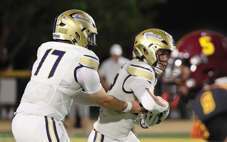 Two people wearing gold helmets and white football uniforms are engaged in a play on a football field at night. Description generated by AI