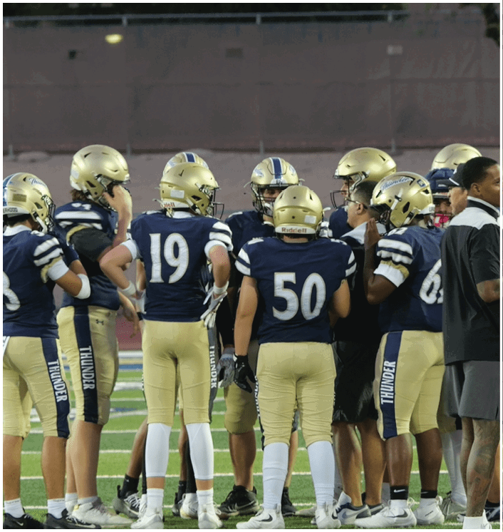 A group of people wearing football helmets and uniforms are gathered on a field with one person in a black shirt standing nearby. Description generated by AI