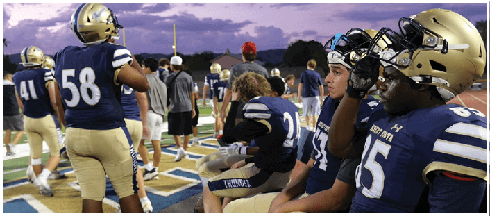 People wearing football uniforms are sitting on a bench on the sidelines of a field. Description generated by AI