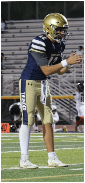 A person wearing a gold helmet and blue and gold football uniform with the word “THUNDER" on the pants is standing on a football field preparing to receive or throw a ball. Description generated by AI