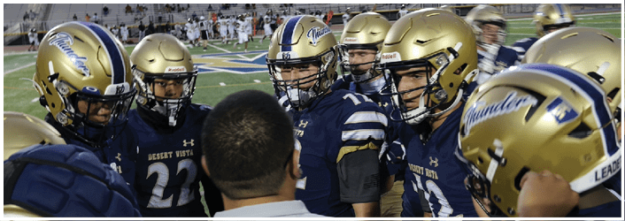 A group of people wearing gold and blue football helmets and uniforms are gathered closely on a field with a stadium and spectators in the background. Description generated by AI