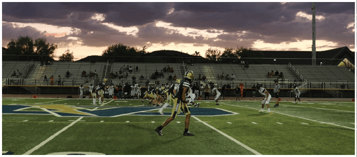 People in sports uniforms are positioned on a football field during a game at dusk with spectators in the stands. Description generated by AI