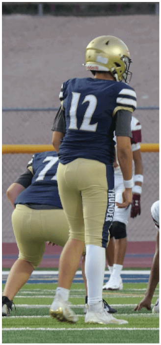 A person wearing a gold helmet and navy blue football uniform with the number 12 stands on a football field preparing for a play while other players in similar uniforms are positioned nearby. Description generated by AI