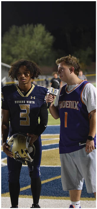A person in a dark football uniform holding a helmet stands on a field while another person in a sports jersey holds a microphone toward them. Description generated by AI