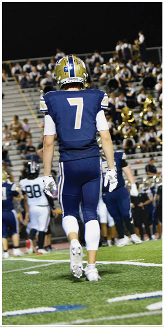 A person wearing a blue football uniform with the number seven on the back is walking on a football field toward a crowd of people and a marching band in the bleachers at night. Description generated by AI