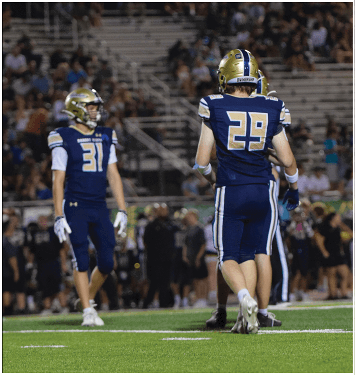 A person wearing a blue and gold football uniform and helmet stands on a field with blurred spectators in the background. Description generated by AI