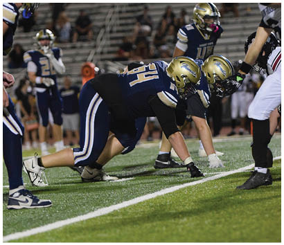 Two football players in blue and gold uniforms are crouched on the field facing an opponent in white pants and black cleats during a game. Description generated by AI