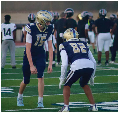 Two football players in navy and gold uniforms are positioned on a field with other players and a coach in the background. Description generated by AI