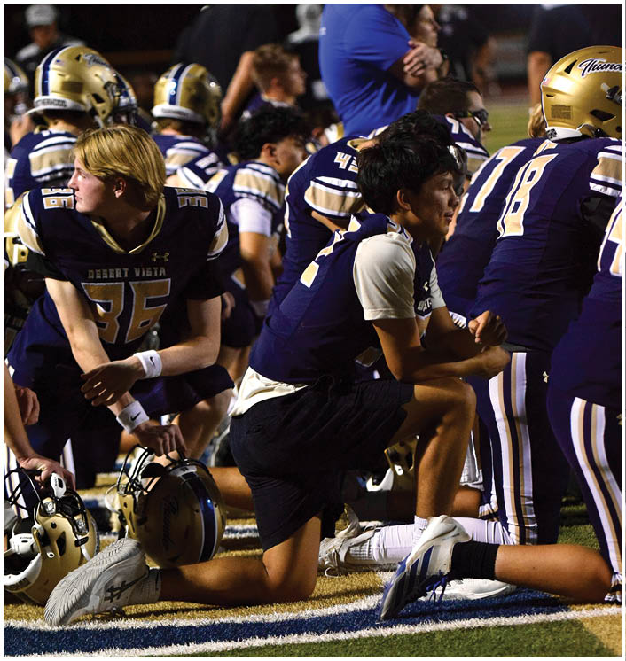 People in football uniforms are sitting and kneeling on a field, some holding helmets, appearing to rest or listen during a game or practice. Description generated by AI