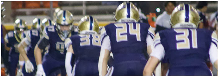 Several people in dark sports uniforms with white gloves are lined up in a crouched position on a field, likely preparing for a play in a football game. Description generated by AI