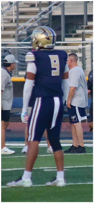 A person in a dark sports uniform with the number nine stands on a football field while two adults in casual athletic clothing stand near the sideline with bleachers in the background. Description generated by AI