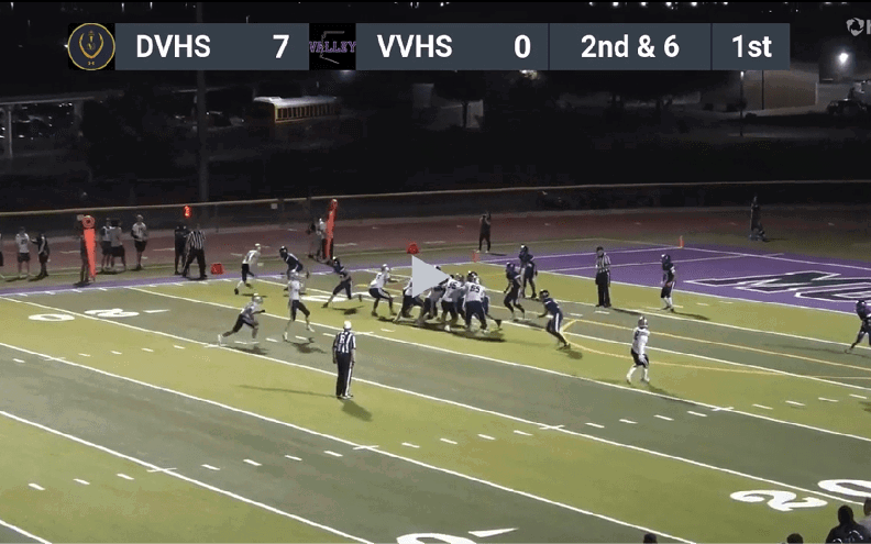 A nighttime football game is in progress on a field with one team in dark uniforms and the other in light uniforms, with the scoreboard showing one team leading and the current down and distance displayed. Description generated by AI