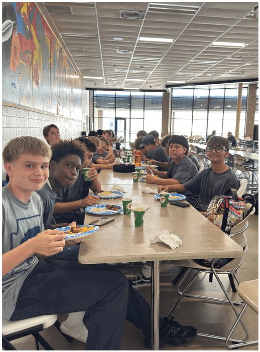 People sitting closely together at a long cafeteria table eating and drinking in a brightly lit room with large windows. Description generated by AI