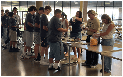 People stand in line at a table indoors where others are serving food and handing out items. Description generated by AI