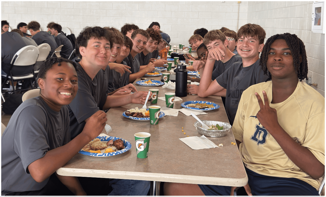 A group of people wearing casual clothing are sitting closely together at a long table in a cafeteria, smiling and eating food from plates with cups and utensils on the table. Description generated by AI