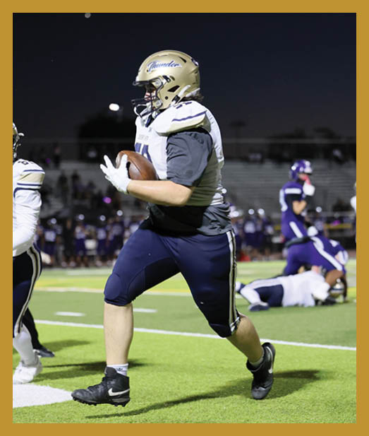 A person in sports gear is running with a football on a field while other players and a referee are visible in the background. Description generated by AI