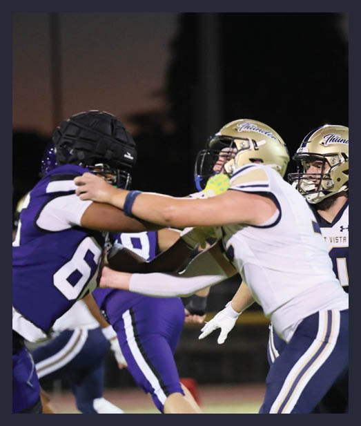 Two people in football uniforms and helmets are engaged in a physical play on a field during a game at dusk. Description generated by AI