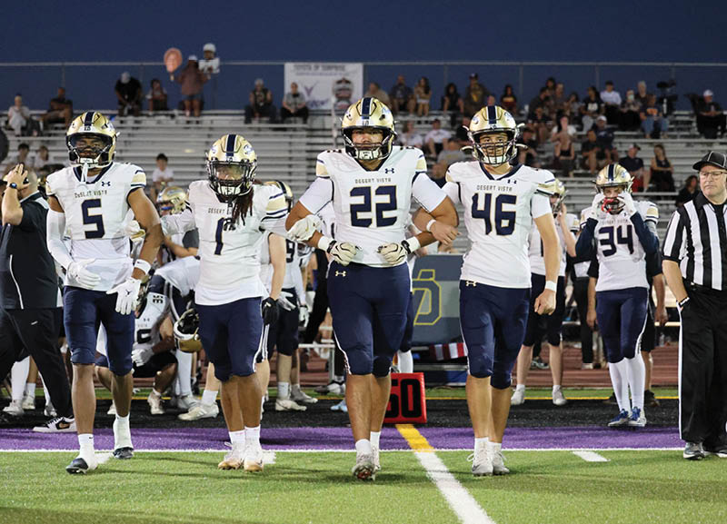 Four people in football uniforms walk arm in arm on a field with a crowd in the background during a nighttime game. Description generated by AI